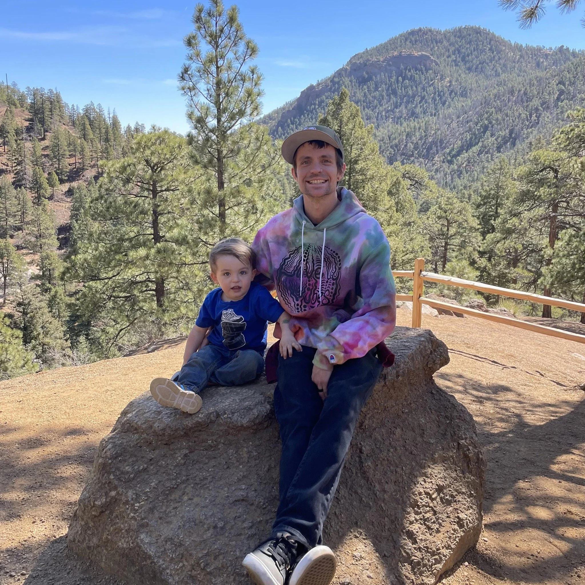 A man and a young boy sitting on a large rock in a forested mountain area with trees and a mountain in the background. The man is smiling, wearing a tie-dye hoodie and a cap, and the boy is looking at the camera, wearing a blue t-shirt and jeans.