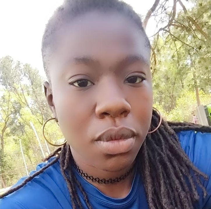 Close-up selfie of a young woman with dark skin and dreadlocks, wearing large hoop earrings, a black choker, and a blue shirt, outdoors with trees in the background.