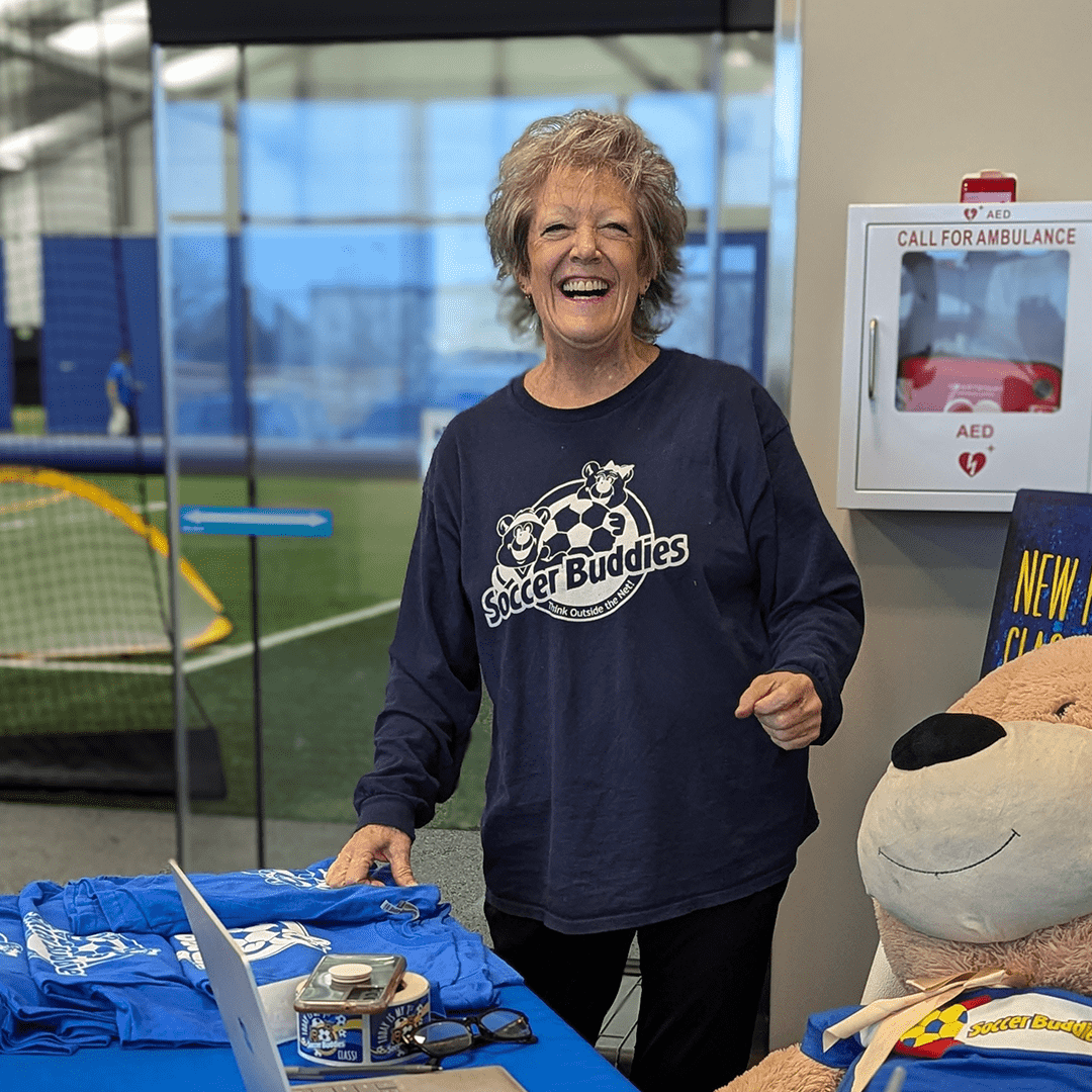 An elderly woman smiling at a table with soccer merchandise at an indoor soccer facility