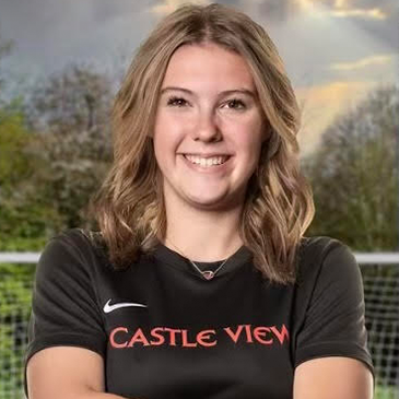 Smiling young woman with wavy blonde hair wearing a black sports shirt with 'CASTLE VIEW' on it, outdoors with trees and a cloudy sky in the background.
