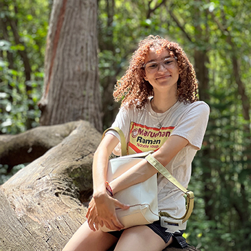 Young woman with curly hair, glasses, and a casual t-shirt sitting in a forest, leaning on a fallen tree trunk, smiling at the camera.