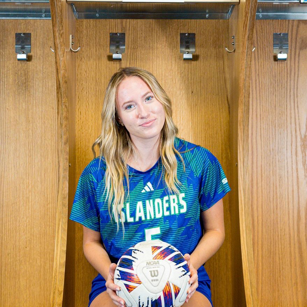 A young woman with long blonde hair holds a volleyball while sitting in a wooden locker room. She wears a blue sports jersey with 'ISLANDERS' written across the front and the number 5.