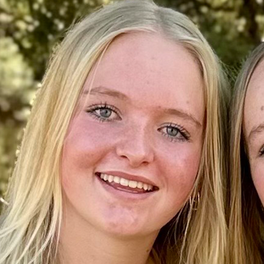 Close-up of a young woman with blonde hair and blue eyes smiling outdoors.
