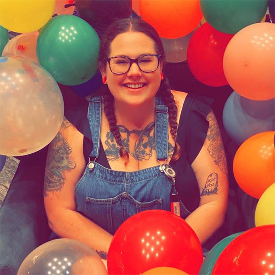 A smiling woman with glasses, dark hair in braids, wearing denim overalls, surrounded by colorful balloons.
