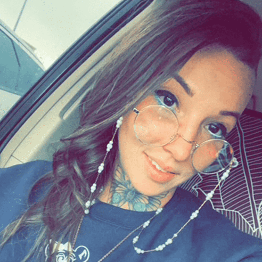 A woman with long wavy hair, wearing round glasses with a pearl chain, smiling, in a car, with a tattoo on her neck and a striped shirt in the background.