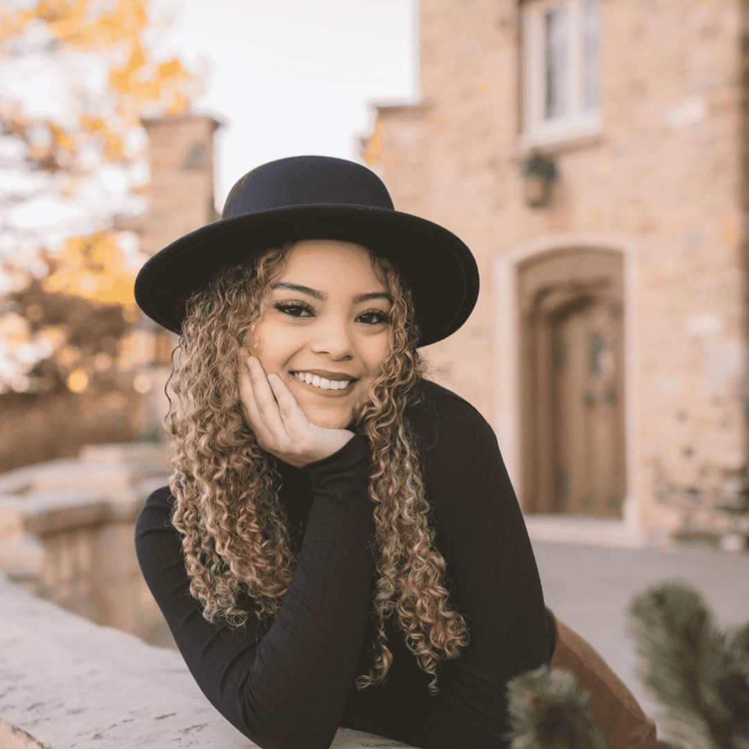 A woman with curly hair wearing a black hat and black top, smiling outdoors in front of a brick building.