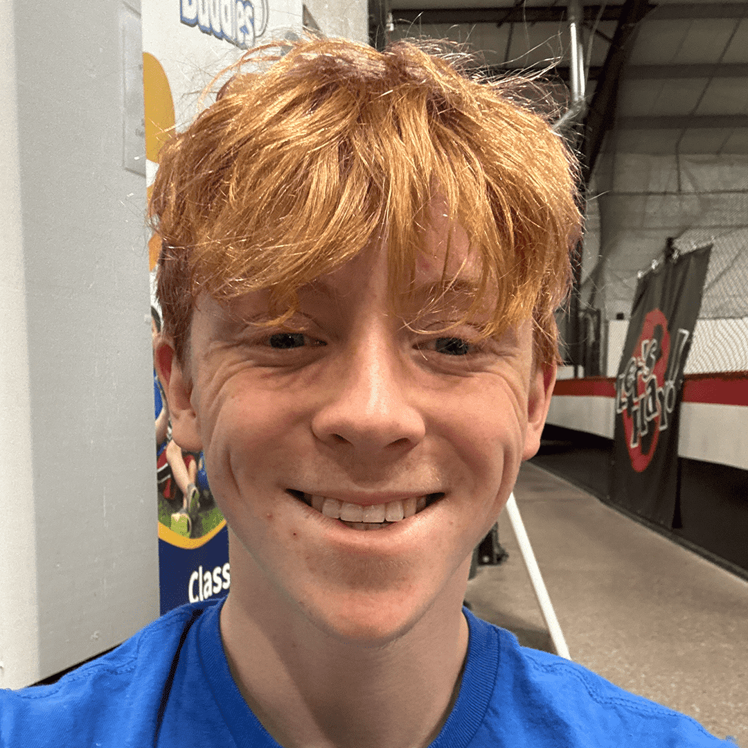 A young man with red hair smiling in an indoor skate park or sports facility.