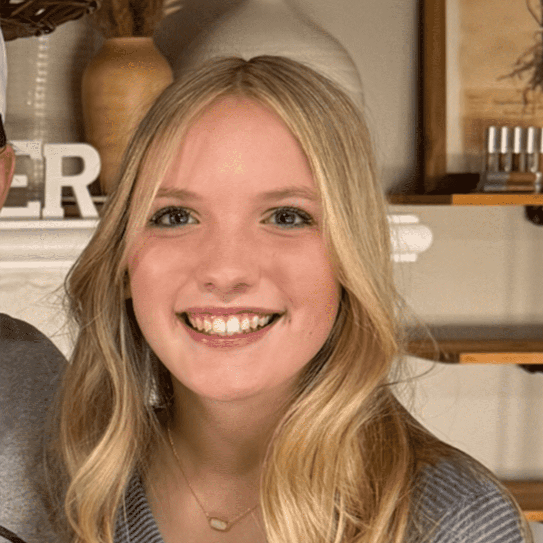 A young woman with long blonde hair smiling, wearing a striped shirt and a small necklace, indoors with wooden shelves and decorative items in the background.