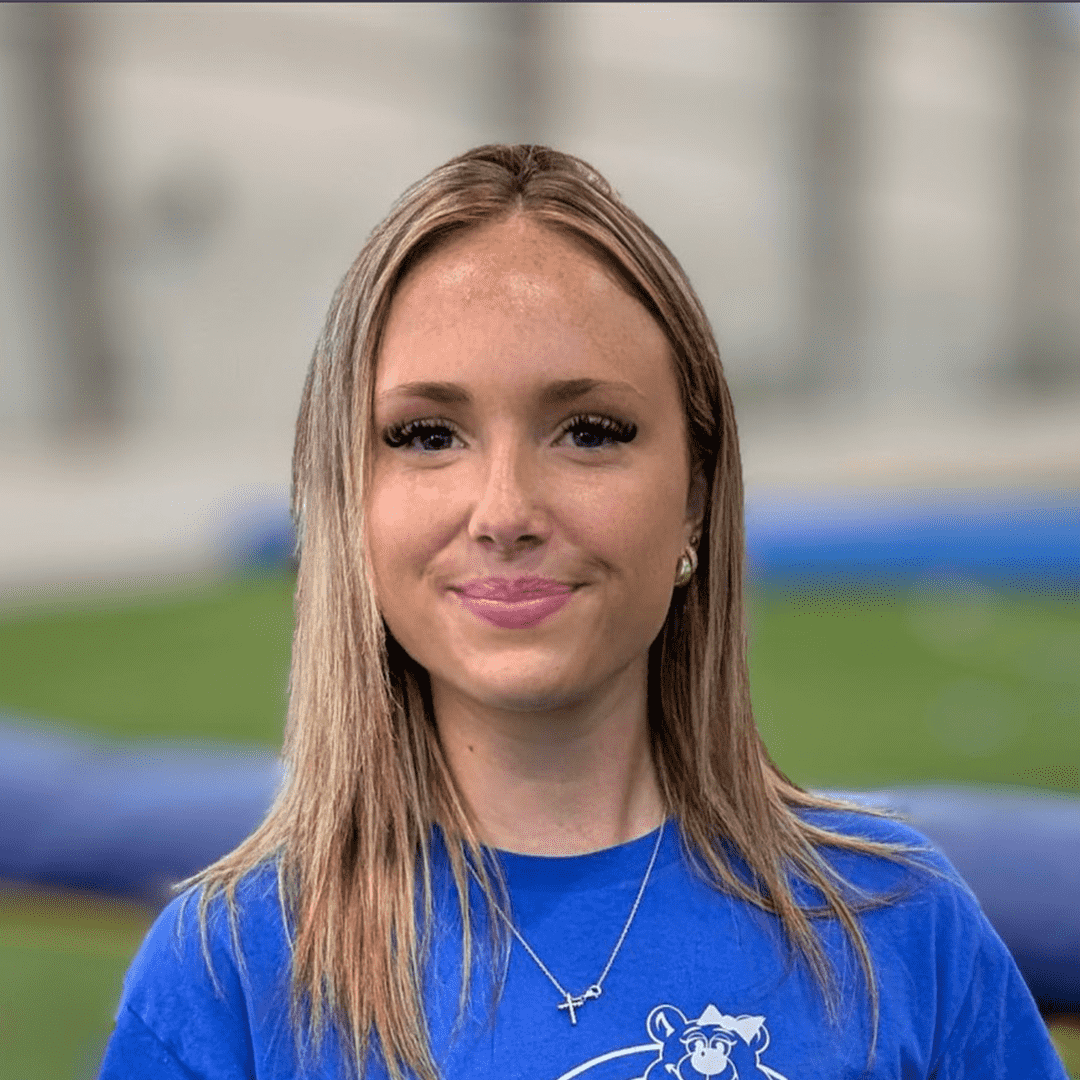 A young woman with long brown hair and makeup, smiling, wearing a blue shirt with a logo of a bear and a cross necklace, outdoors with a blurred background.