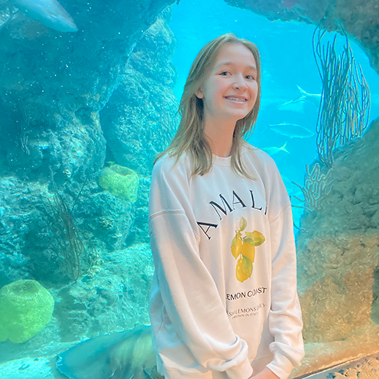 A girl with long blonde hair smiling at an underwater exhibit, surrounded by coral and fish.