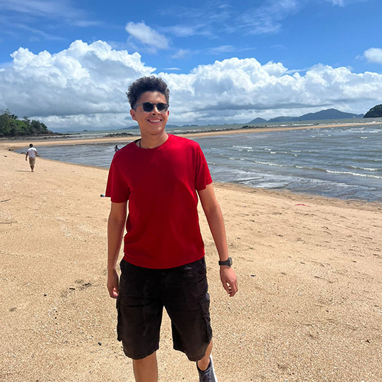 A man wearing sunglasses, a red t-shirt, and black shorts walking on a sandy beach with ocean waves, cloudy sky, and distant mountains in the background.