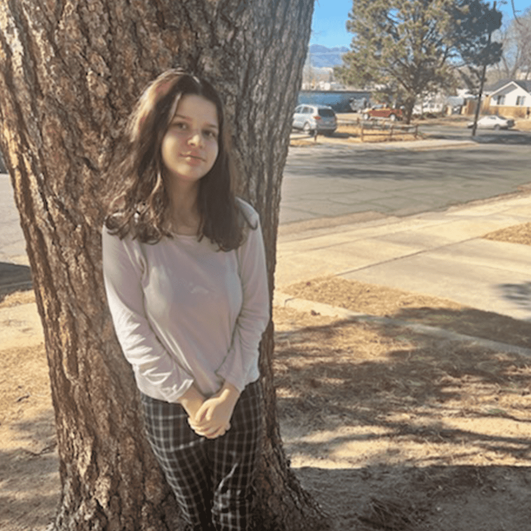 A young woman with shoulder-length dark hair stands leaning against a large tree trunk with both hands clasped in front of her. She wears a light-colored long-sleeve top and checkered pants. The background shows a suburban neighborhood with parked cars, a sidewalk, and trees, under a bright, clear sky.