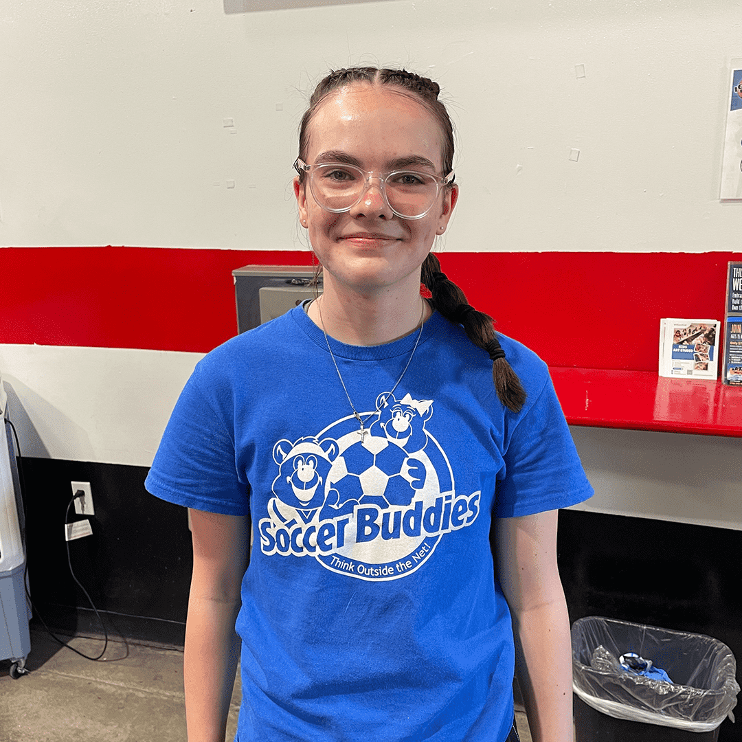 A young girl with glasses in a blue Soccer Buddies t-shirt standing indoors near a counter with pamphlets and a trash bin.