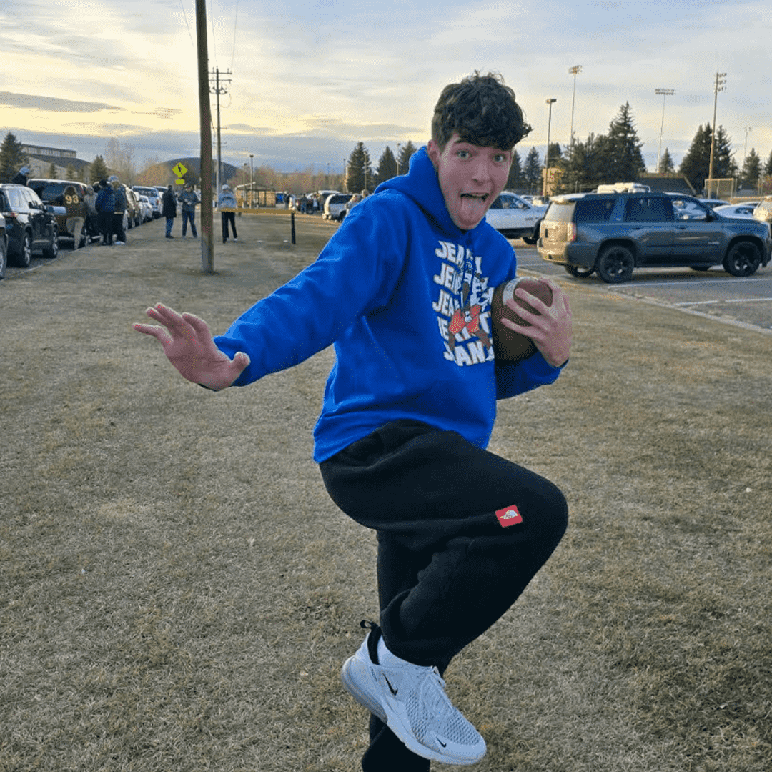 A young man in a blue hoodie and black sweatpants balancing on one leg outdoors while holding a football, with a surprised or playful expression. In the background, there are parked cars, a parking lot, and a few people near a sports field under a partly cloudy sky.