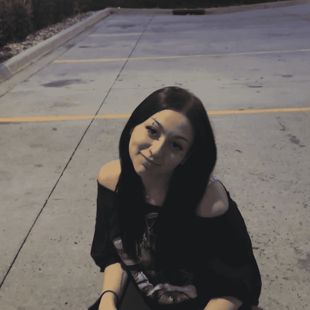 A young woman with long dark hair sitting on the concrete ground outside at night, smiling at the camera, wearing a black off-shoulder top.