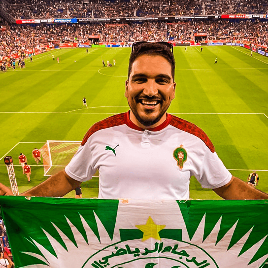 A man smiling on a soccer field holding a green and white flag with Arabic writing, wearing a Moroccan soccer jersey, with a large crowd in the stadium behind him.
