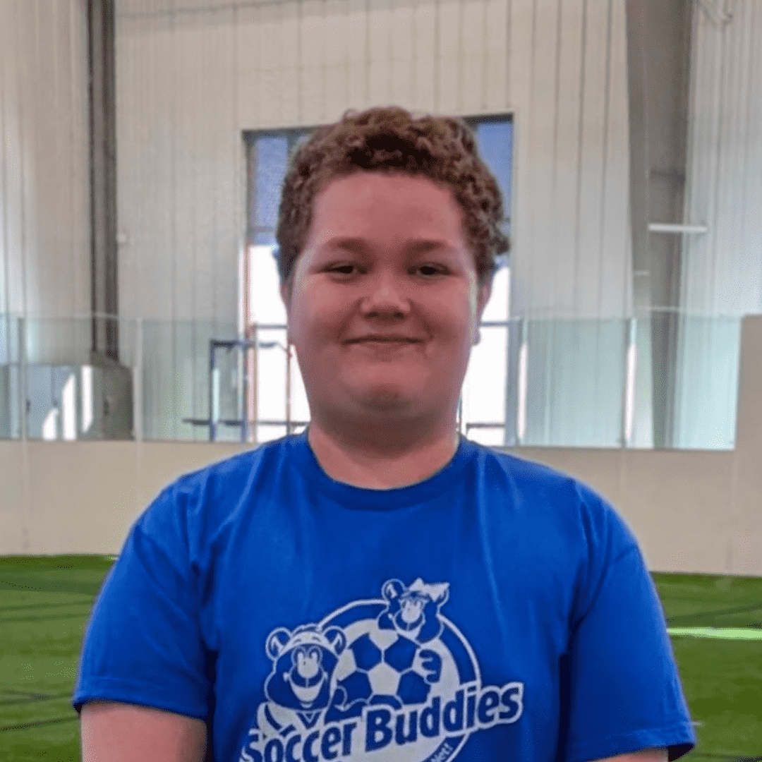 A young person with short curly hair smiling, wearing a blue T-shirt with a Soccer Buddies logo, in a sports facility.
