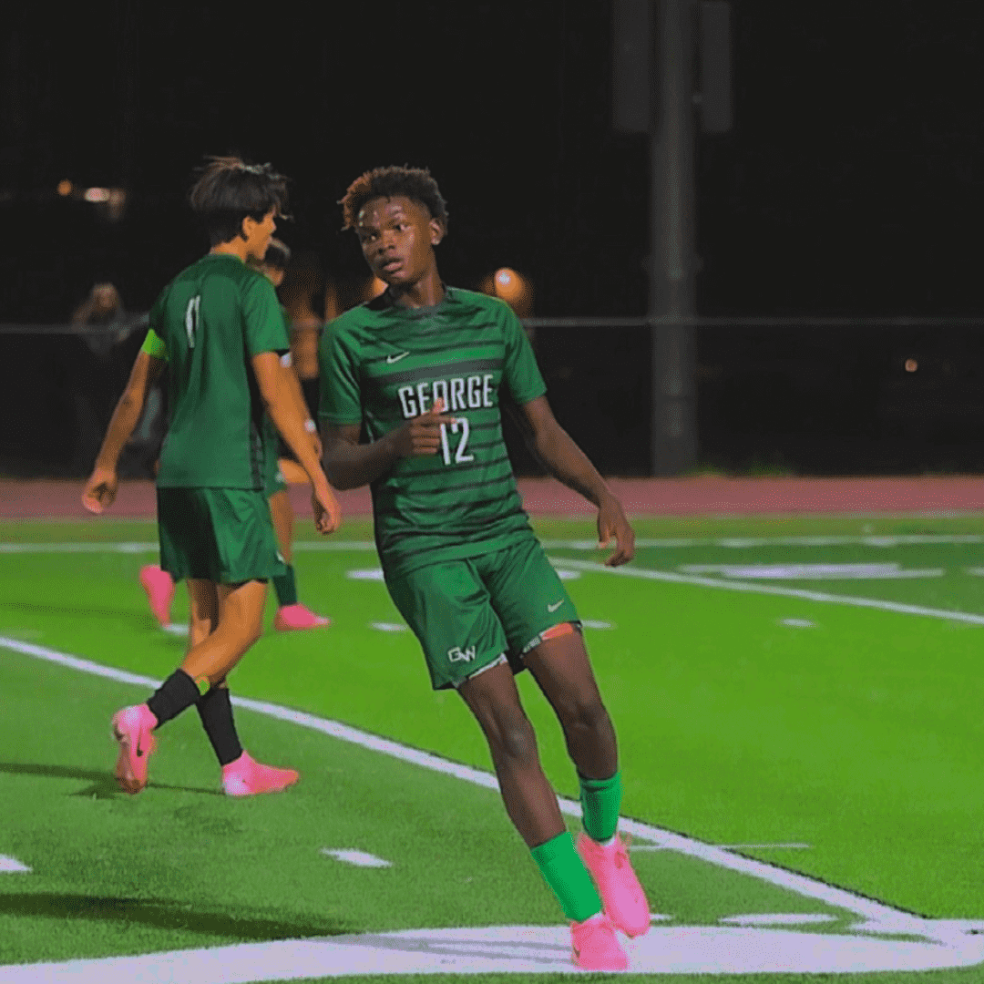 Young soccer player in green uniform running on a soccer field at night.