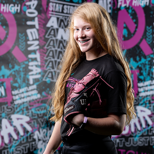 A young girl dressed in a black sports t-shirt and pink baseball glove standing in front of a colorful, graffiti-style wall with motivational words and phrases related to sports and perseverance.