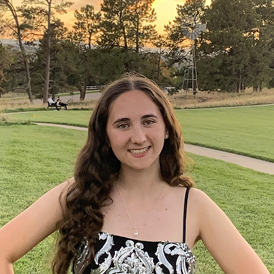Young woman with long curly brown hair smiling outdoors in a park at sunset, wearing a black and white patterned dress and a necklace.