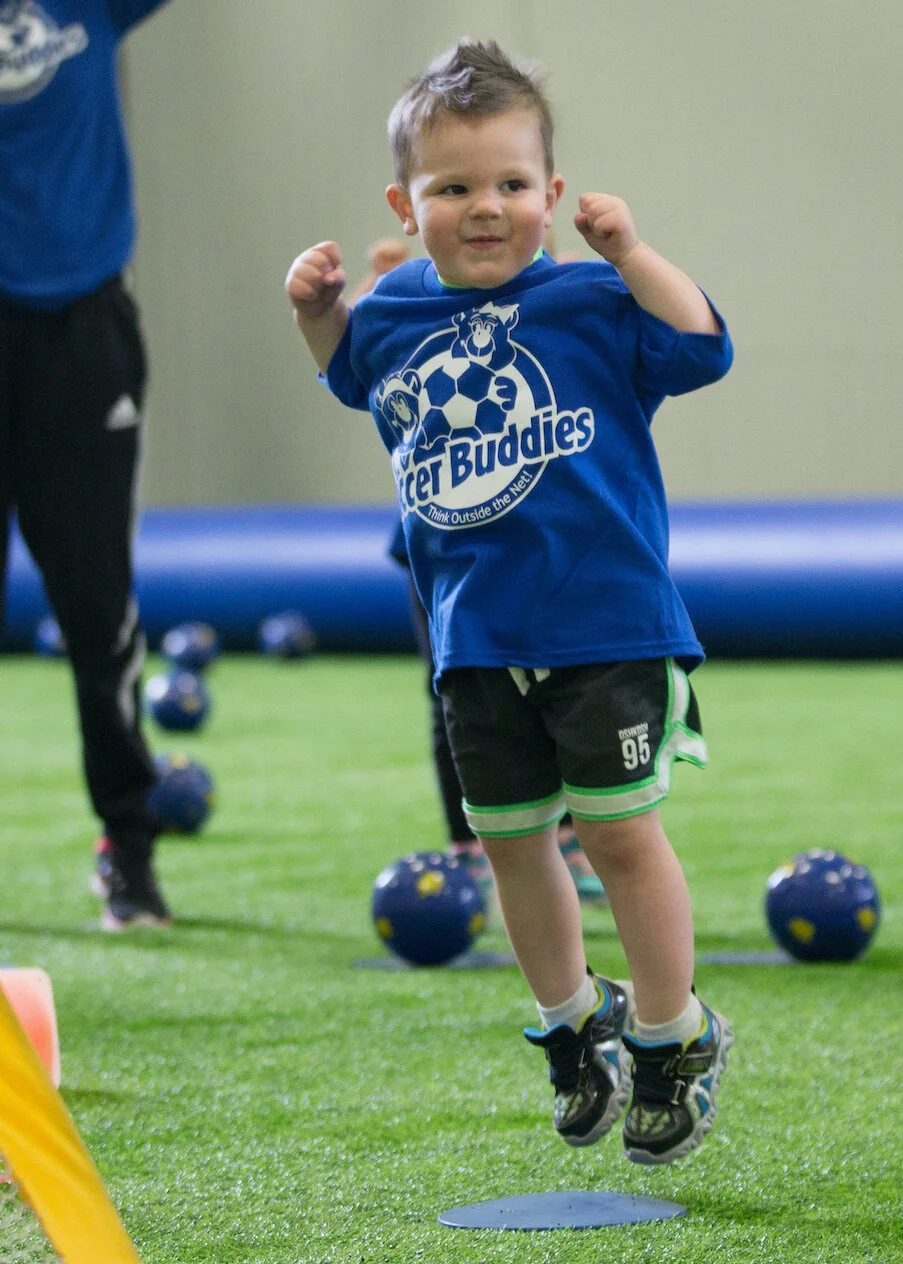 A young boy jumping with a big smile, wearing a blue 'Soccer Buddies' t-shirt, black shorts with green trim, and sneakers, in an indoor sports facility with exercise balls and a green artificial turf floor.