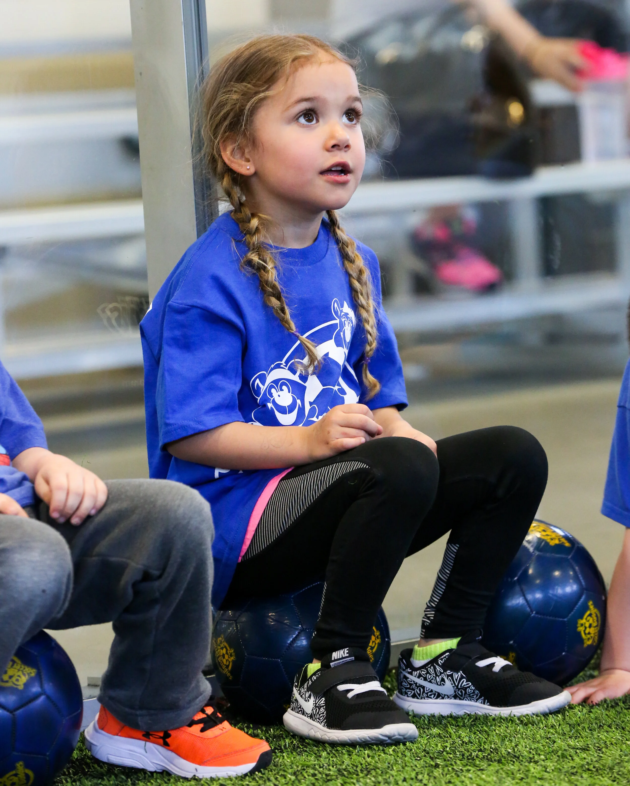 A young girl with braided hair sitting on a soccer ball inside a sports facility. She is wearing a blue sports shirt, black leggings, and black and white athletic shoes. She appears to be attentively watching or listening to something.