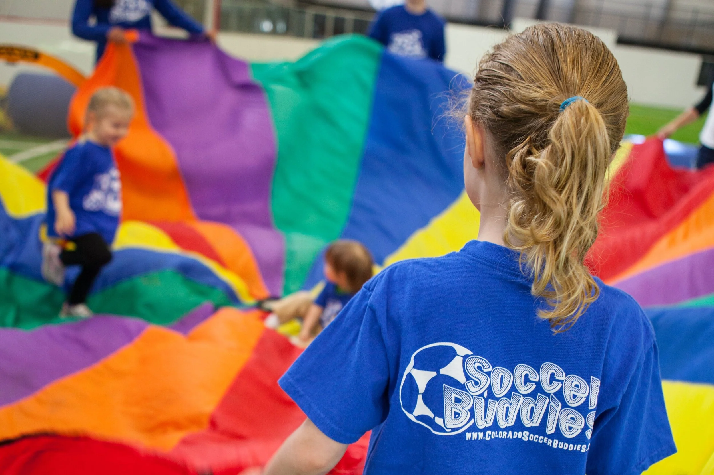 Children playing on a colorful parachute indoors, with one girl in a blue shirt with 'Soccer Buddies' logo in the foreground.