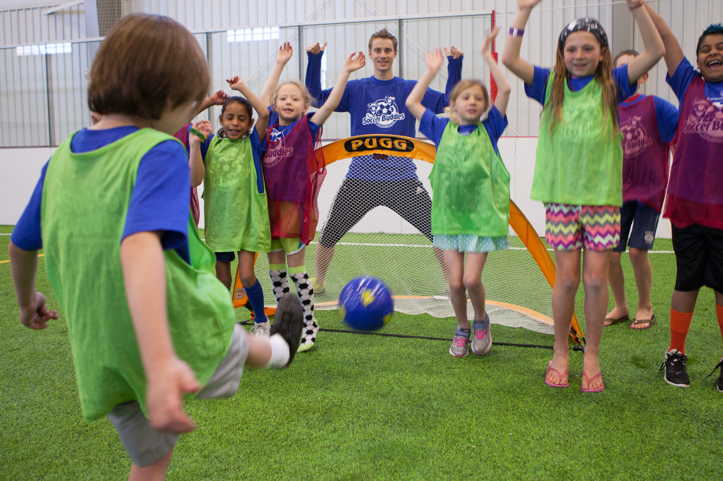 Children playing soccer indoors, with one child kicking a blue ball towards a goal, while others watch and cheer.