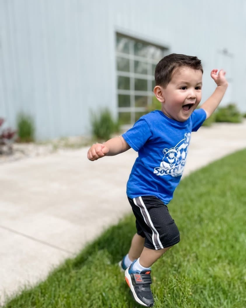 Child running and celebrating outdoors on a sidewalk next to grass, wearing a blue t-shirt, black shorts, and sneakers.