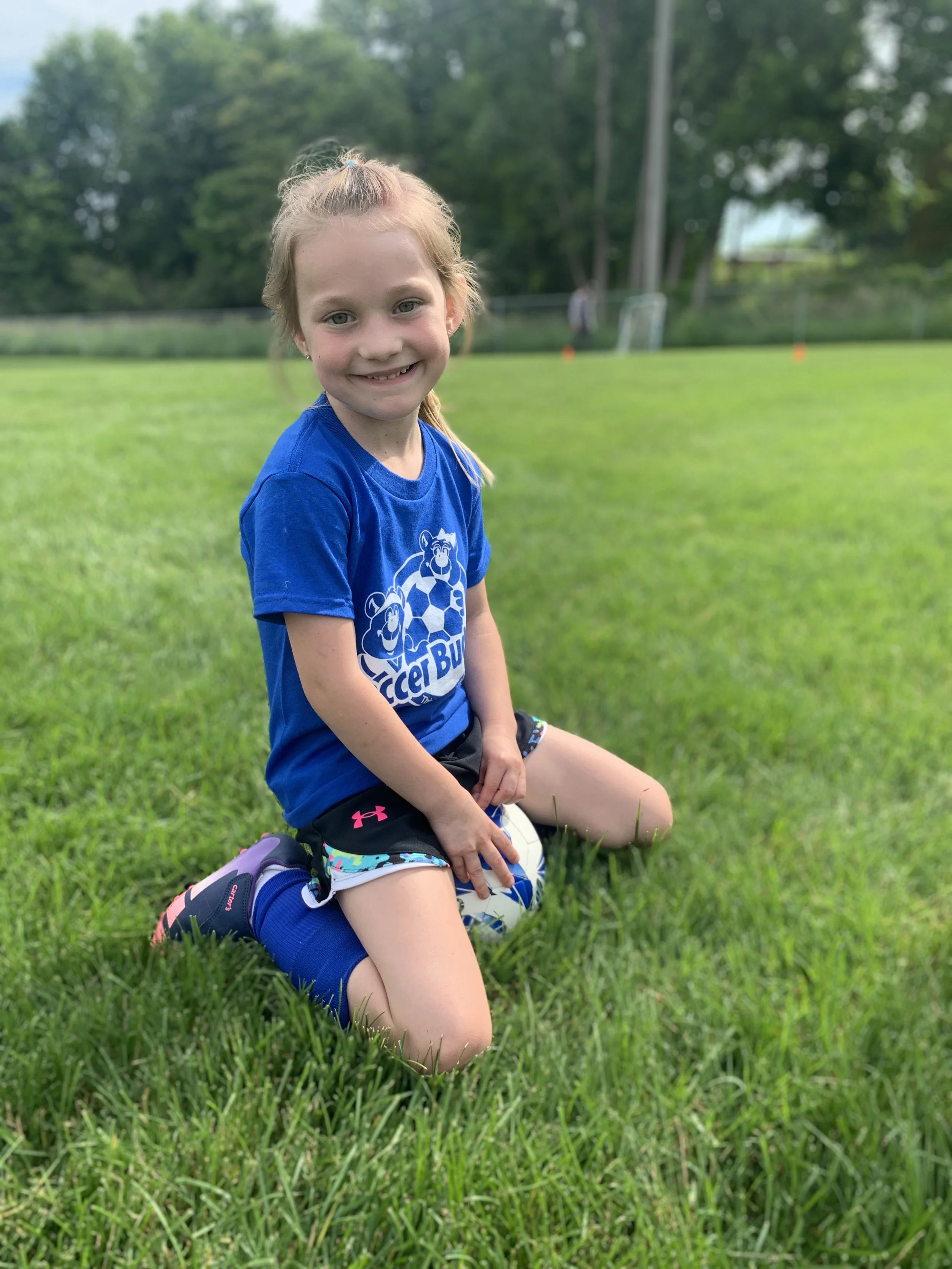 Young girl kneeling on grass holding a soccer ball, smiling, in a park or sports field.