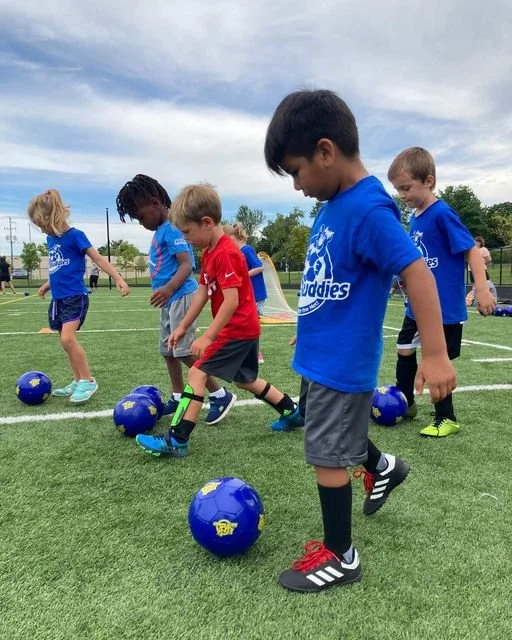 Young children practicing soccer skills on a field with blue soccer balls, some wearing blue shirts, one in red, under a partly cloudy sky.