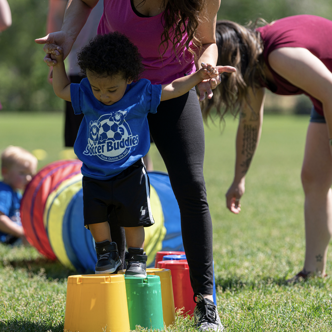 Young boy in a blue shirt balancing on colorful plastic buckets during an outdoor activity, with adults and a play tunnel in the background.