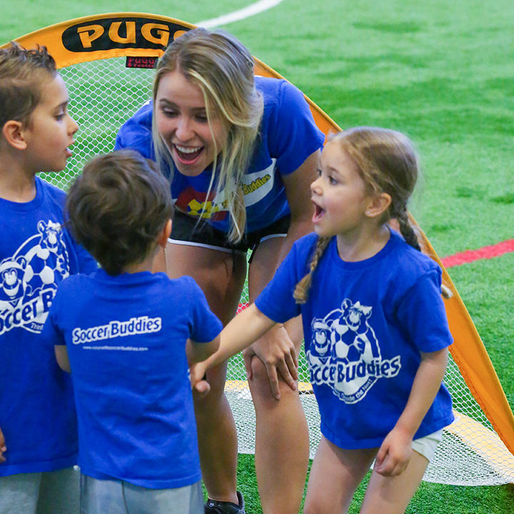 A female soccer coach with blonde hair, wearing a blue shirt, happily interacting with young children in blue Soccer Buddies shirts on a field.