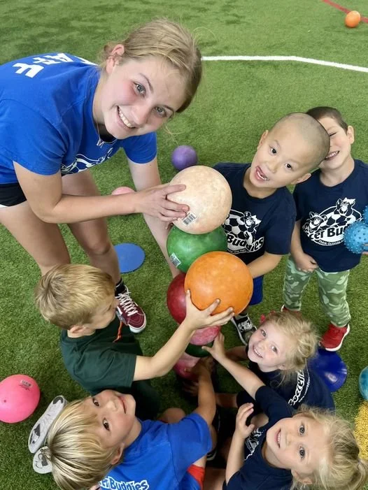 Group of children and a young woman smiling and stacking colorful balls on a grassy field.