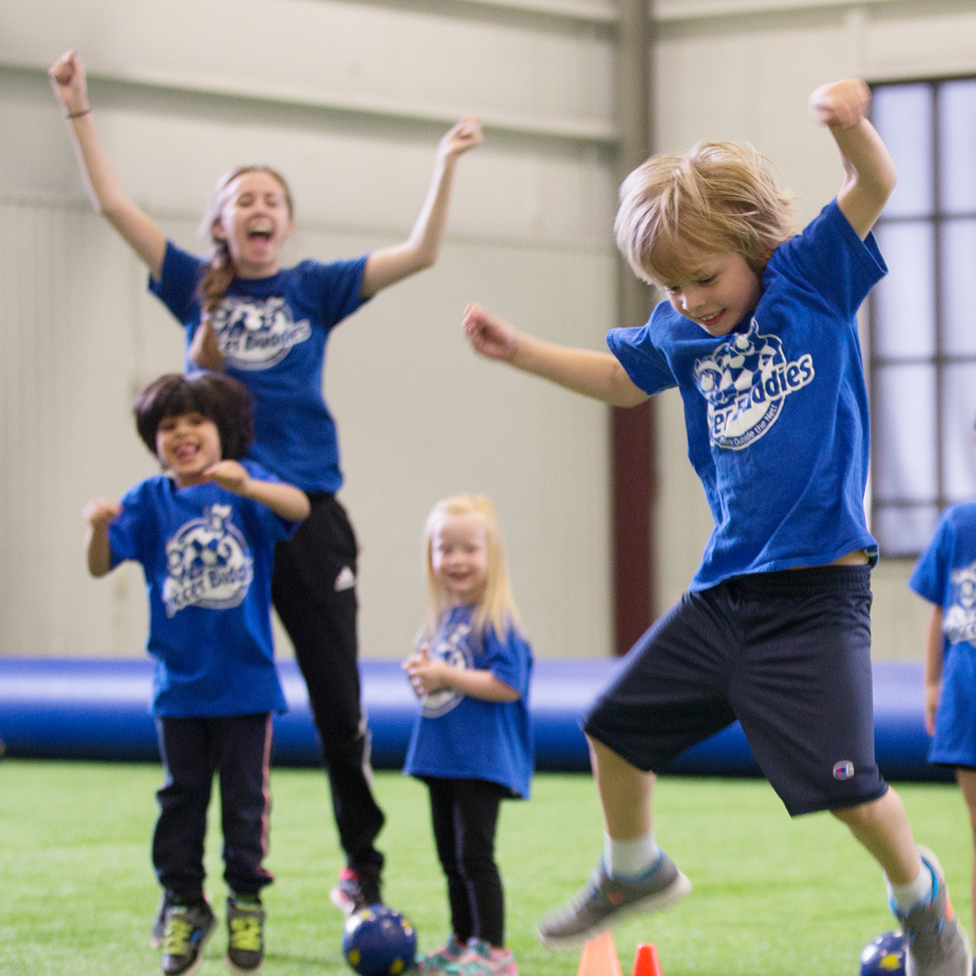 Children and an adult in a gym or indoor sports facility, wearing blue soccer t-shirts, participating in a fun activity or game involving jumping over cones.