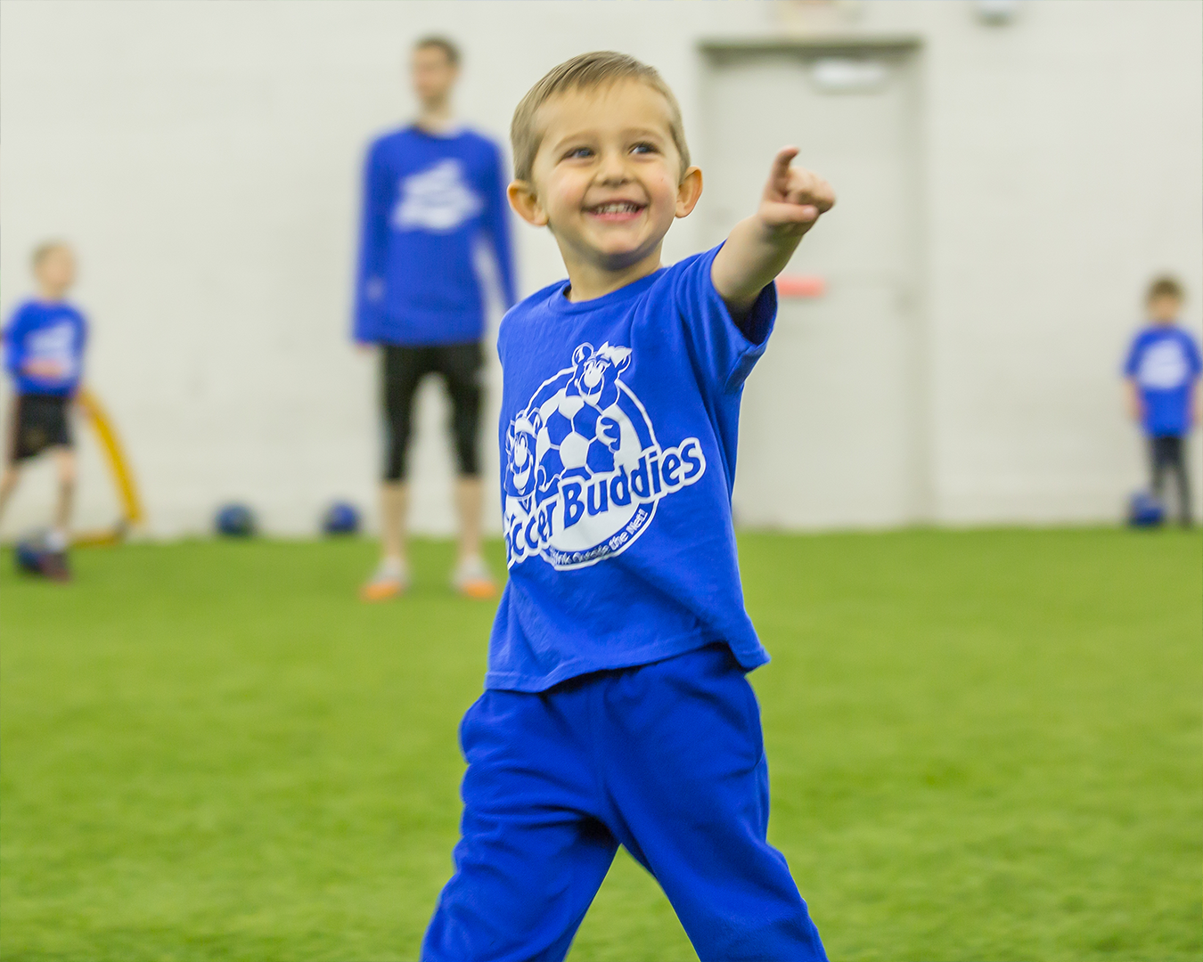 A young boy in a blue soccer uniform pointing and smiling during a soccer practice session, with other children and an adult coach in the background.
