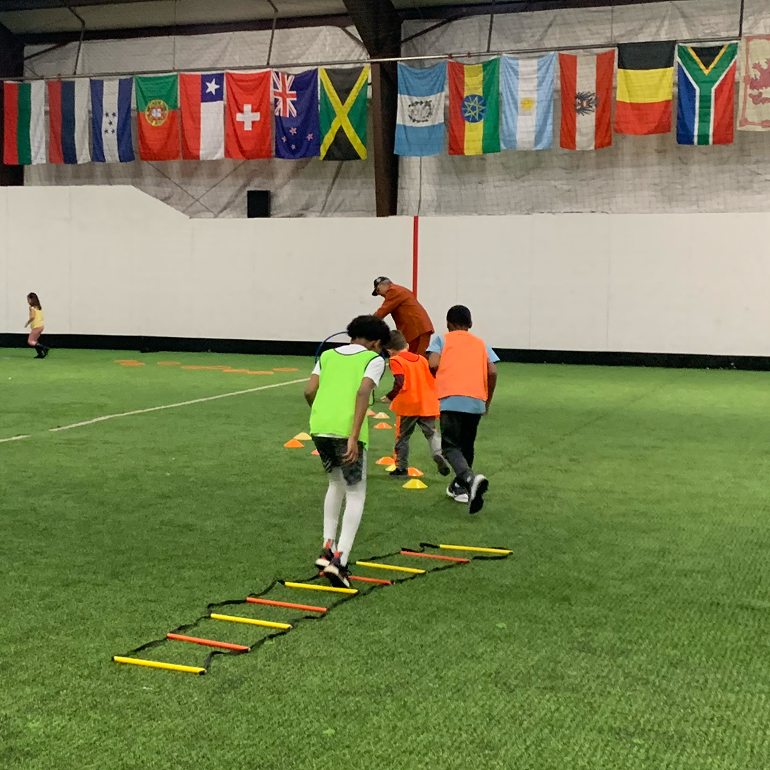 Children participating in an indoor athletic training session, practicing agility drills with hurdles and cones on a green turf field, with international flags hanging above in the background.