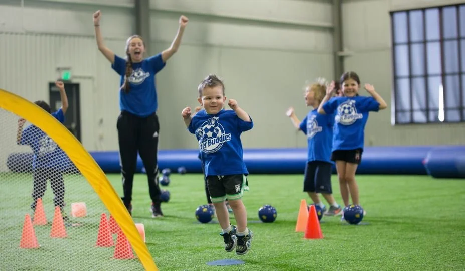 Children and an adult participating in an indoor soccer game, with children jumping and a woman cheering in the background.