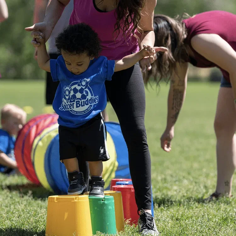 A young boy walks on colored cones during an outdoor activity, with adults assisting and children playing in the background.
