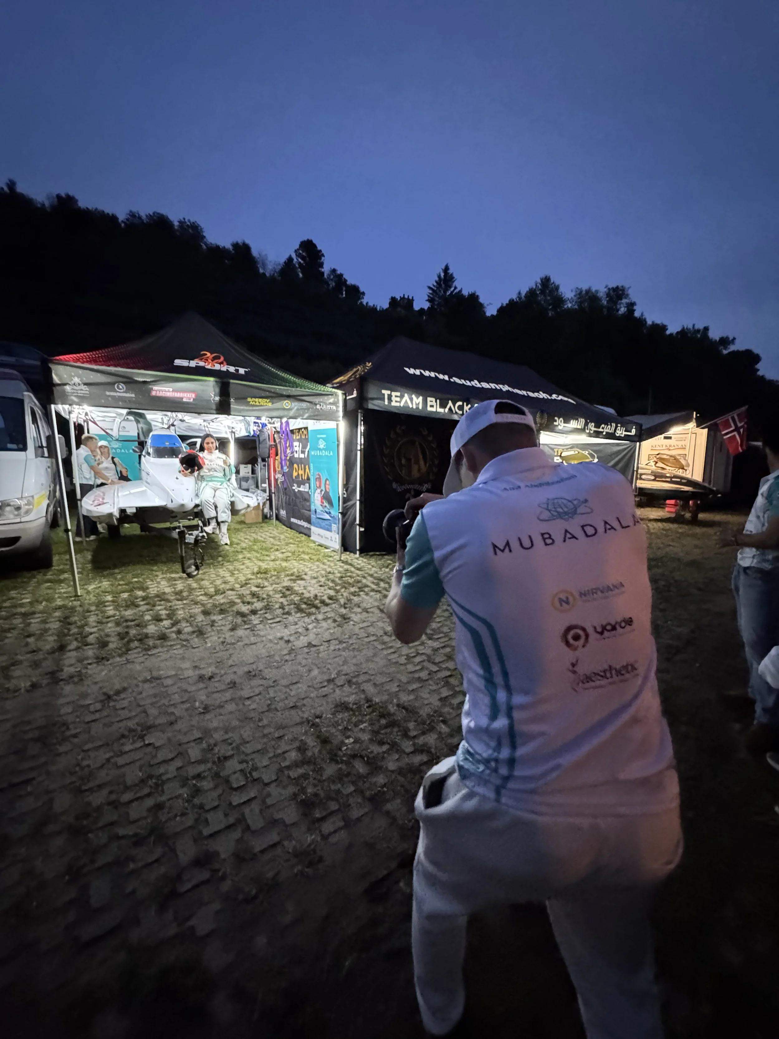 En person i en hvit og blå sportskjorte, sett bakfra, som tar et bilde av en vogn og mennesker under en opplyst campingåpen telt på kveldstid.