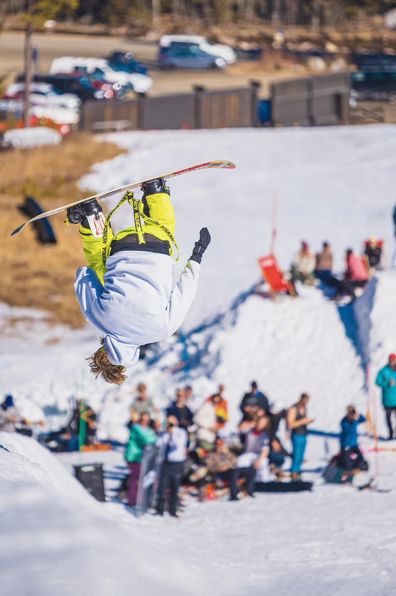 En person som utfører en bakovers Dort i snøen på en snowboarding-konkurranse med tilskuerer og biler i bakgrunnen.