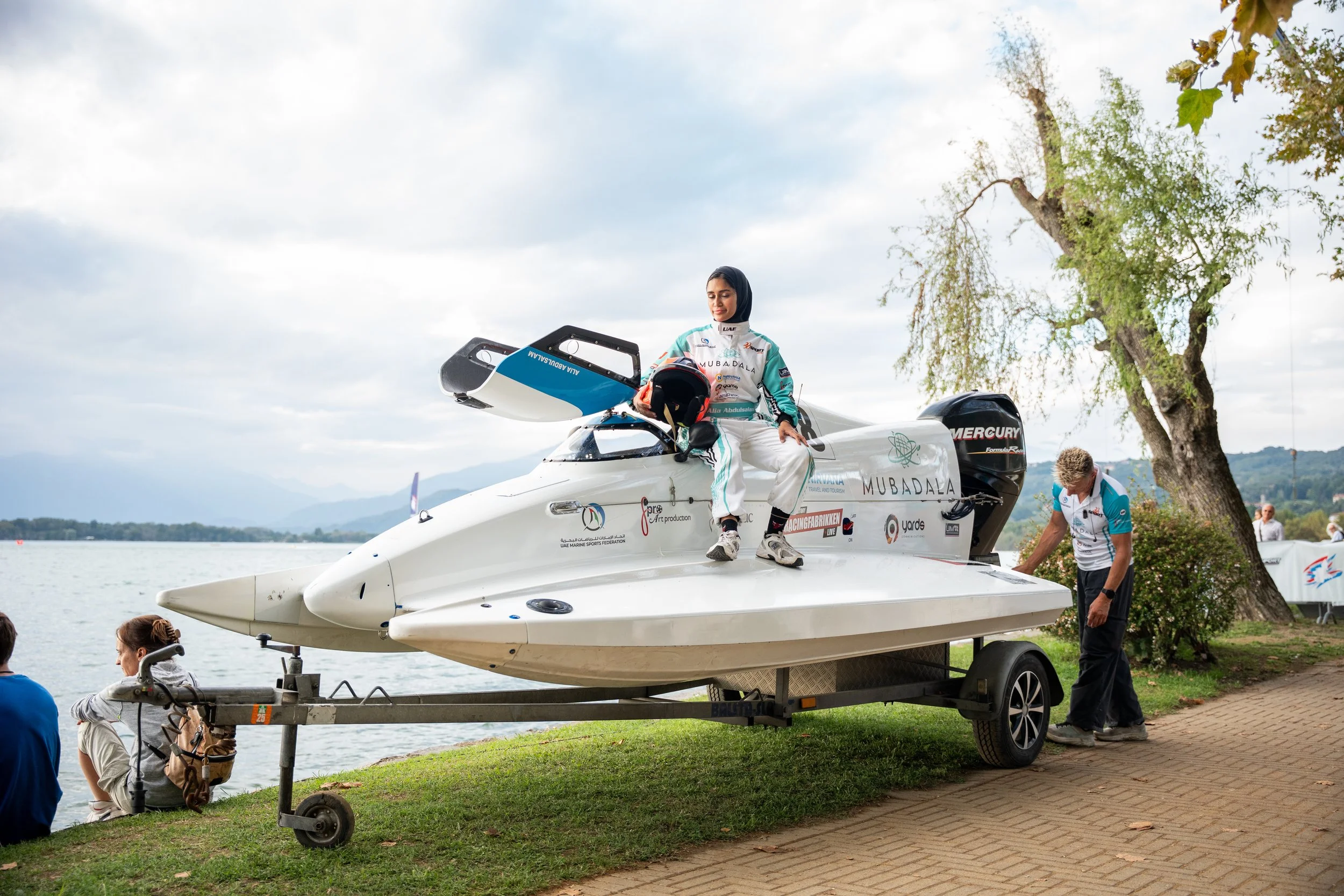Kvinne i racingdrakt sitter på en hurtigbåtmotor à bord av en forhøyning, med vann og fjell i bakgrunnen, mens andre personer står og sitter i nærheten.