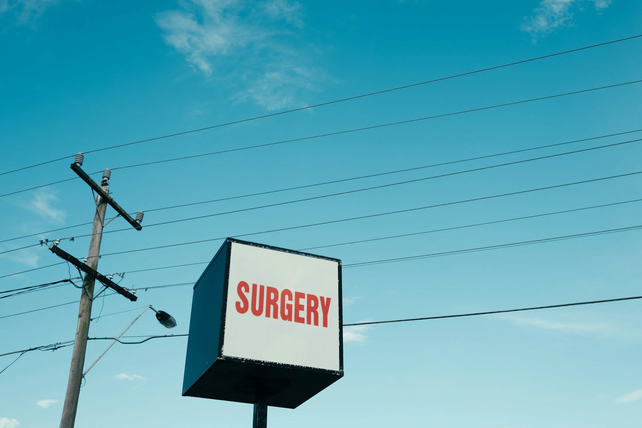 Electric utility pole with multiple power lines under a blue sky with scattered clouds, partially obscured by a sign that reads 'SURGERY' in red letters.
