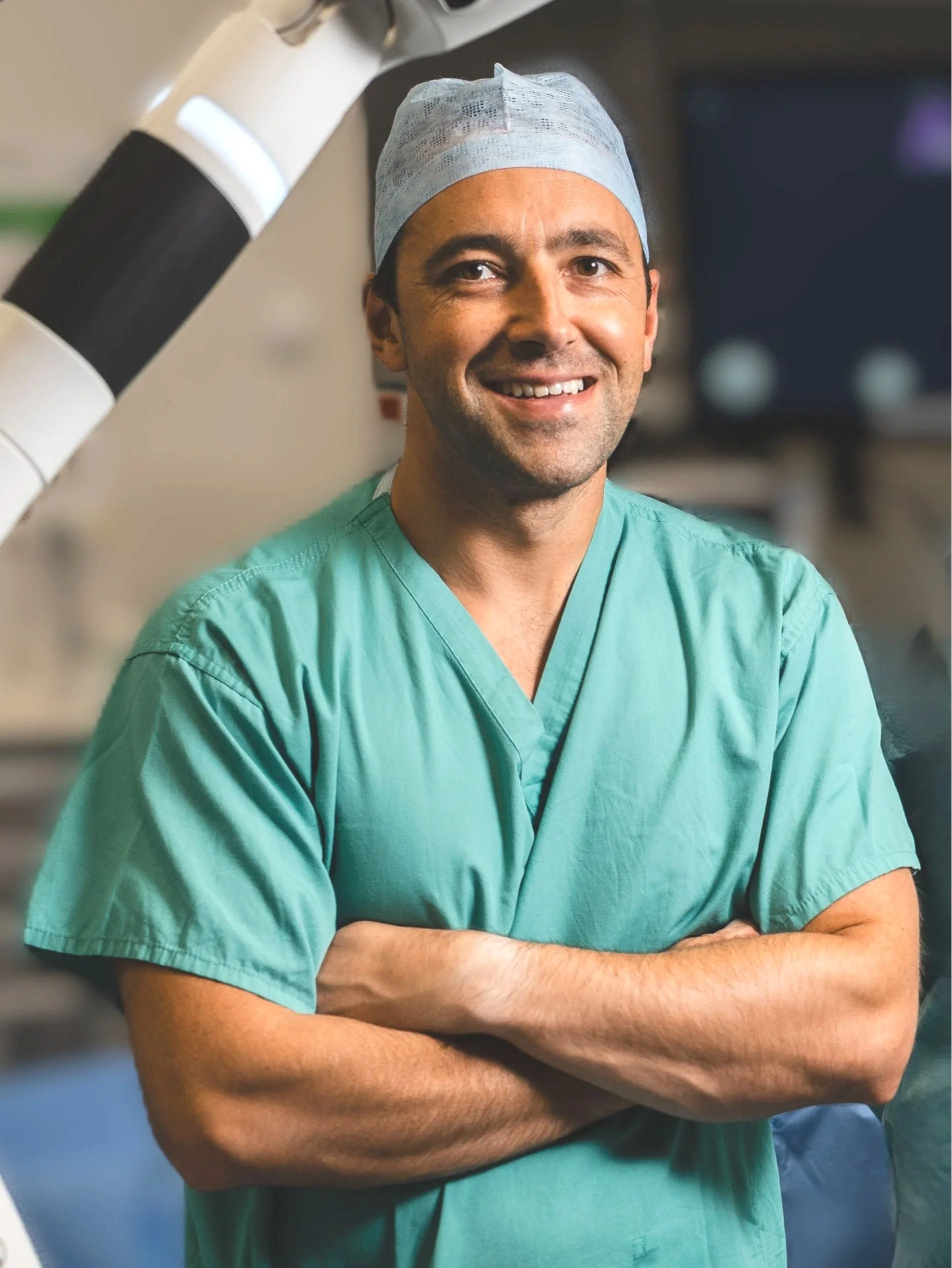 A smiling male doctor or surgeon in green scrubs and a surgical cap, posing with arms crossed in a hospital or operating room.