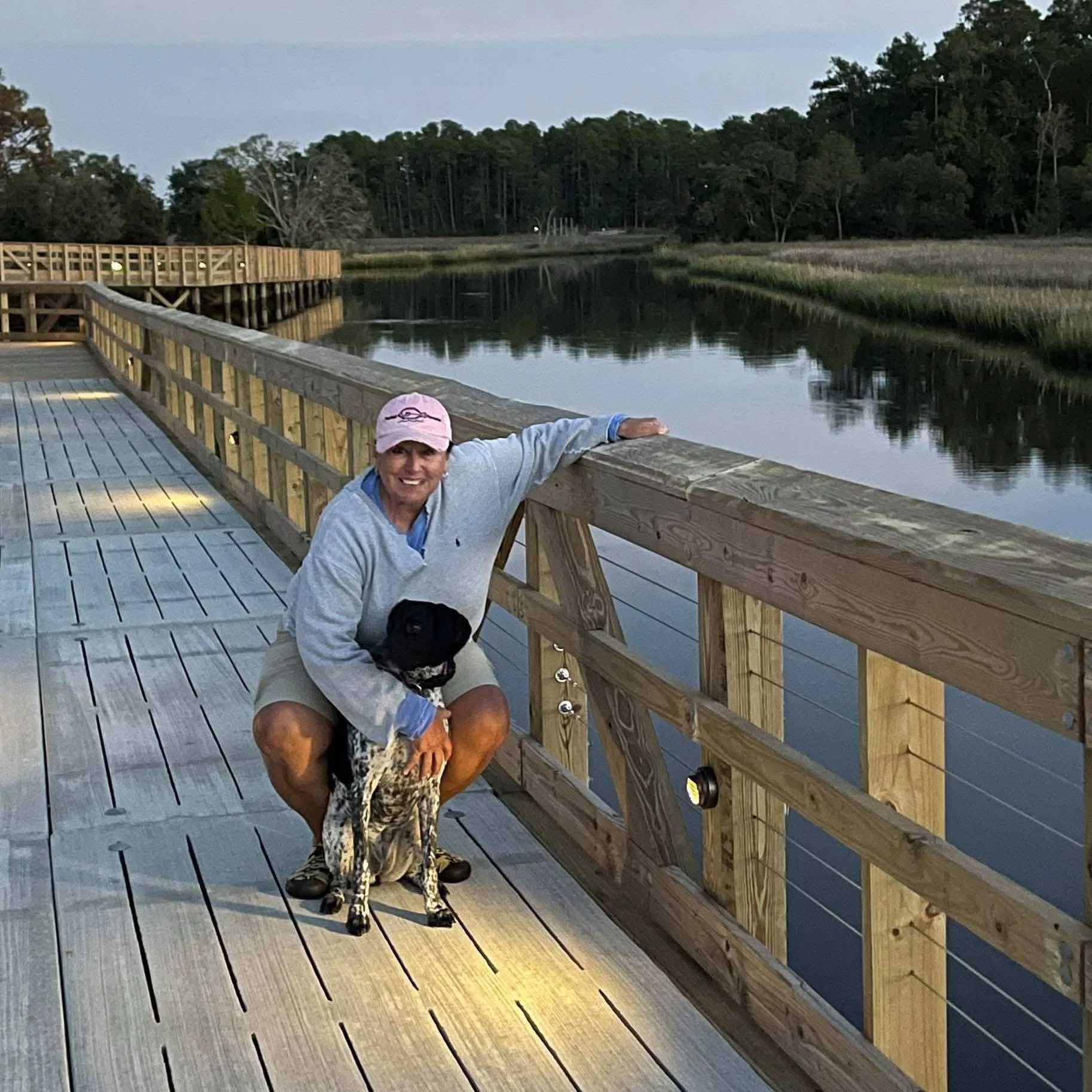 A woman in a pink cap crouching on a wooden dock, holding a black and white dog, with a calm body of water and trees in the background.