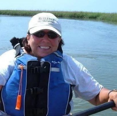 Woman smiling in a kayak on a body of water, wearing a white cap, sunglasses, and a blue life jacket.