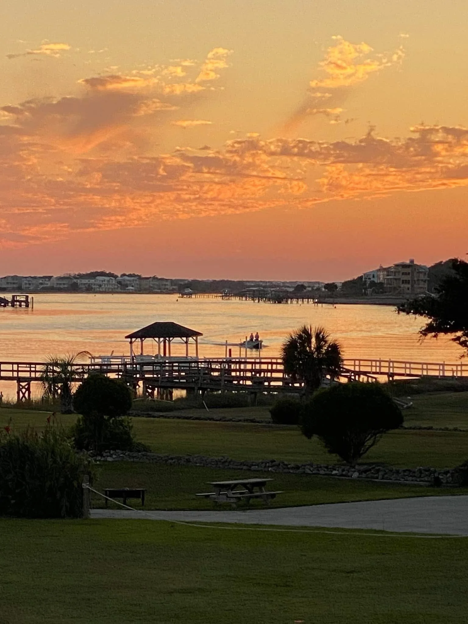 Sunset over a calm body of water, with a wooden pier and gazebo, trees, and a grassy area in the foreground.
