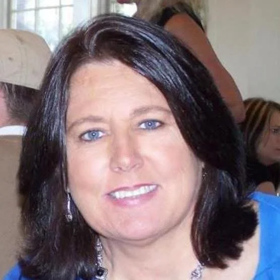 A woman with dark hair and blue eyes smiling at the camera, wearing a blue top and silver jewelry.  Patti Hewett