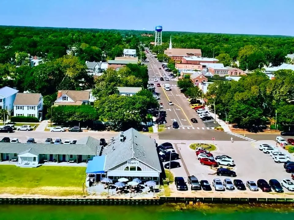 Aerial view of a small town with a main street, parking lots, green trees, and a waterfront restaurant with outdoor seating.