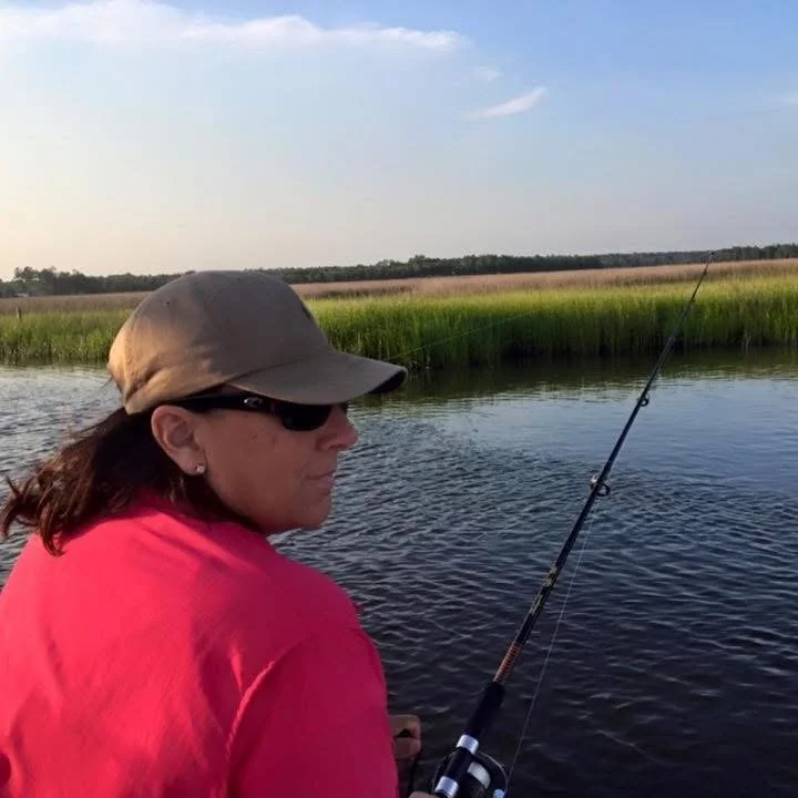Woman fishing on a calm river in a rural area, wearing a baseball cap, sunglasses, and a pink shirt, with grassy fields and trees in the background.